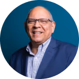 Smiling elderly man wearing glasses, a blue suit jacket, and a checked shirt against a teal background.